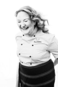 Elizabeth Marshall with her hair down laughing with absolute joy. She's dressed in her chef jacket and half apron. Photo taken by Tabitha Arthur Photography.